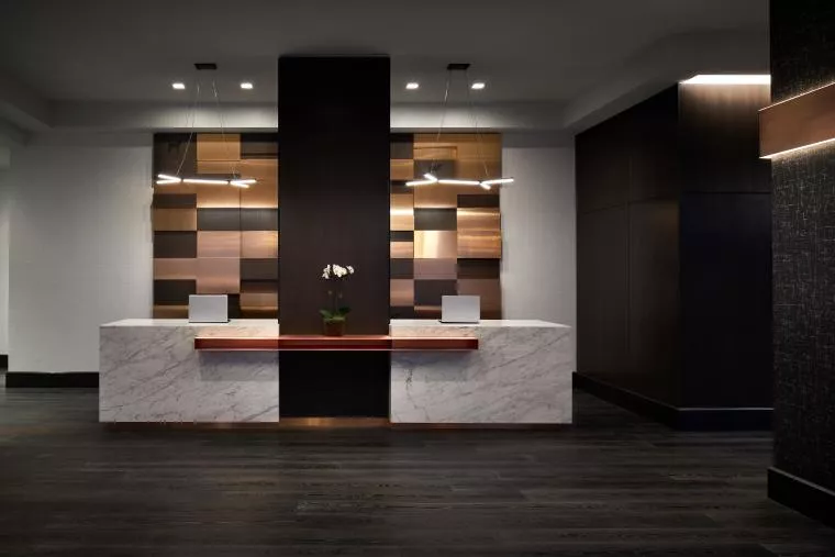 A modern hotel reception desk with two marble counters, each holding a laptop, separated by a central wooden accent wall with geometric wood paneling. A potted orchid sits between the counters. Contemporary pendant lights hang above.