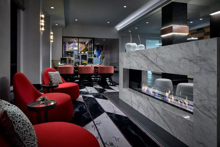 Modern lobby with a marble fireplace, red lounge chairs, small tables, and abstract-patterned carpet. In the background, a dining area with orange chairs and wall art is visible. The space is warmly lit and stylish.