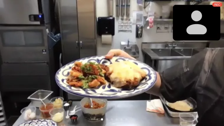 A person in a kitchen holds a plate with two dishes: one is cheesy and baked, the other appears to be pasta with tomato sauce. Various cooking ingredients and utensils are visible on the counter.