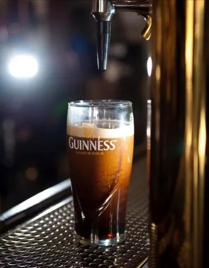 A pint glass of Guinness beer is being filled from a tap, with foam forming on top. The glass sits on a metal bar counter, and bright light is visible in the background.