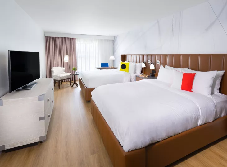 Modern hotel room with two large beds, a flat-screen TV on a light-colored dresser, a white armchair near a window with sheer curtains, and a small table with a lamp and decorative plant.