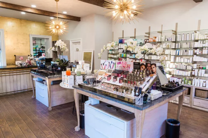 A bright, modern beauty store interior with wooden floors and two display tables filled with makeup products. Shelves with skincare items line the wall, and decorative star-shaped light fixtures hang from the ceiling.