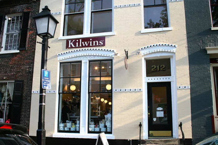 A storefront with cream-colored bricks features a large window displaying treats and a sign reading Kilwins above it. The entrance door is marked with the number 212, and a classic black streetlamp stands on the sidewalk.