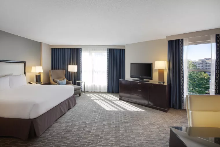 Spacious hotel room with a large bed, armchair, flat-screen TV on a dark wood dresser, two lamps, and large windows with blue curtains letting in natural light.
