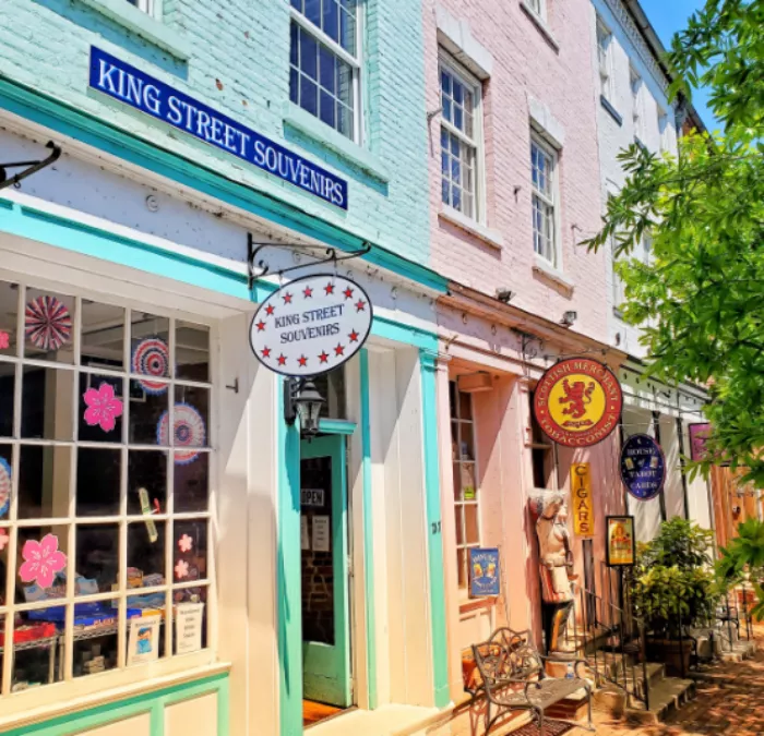 Colorful storefronts with pastel-painted brick walls line a sunny street. Signs for King Street Souvenirs and a cigar shop hang above doors. Large windows display goods, and green leaves frame the right side of the scene.