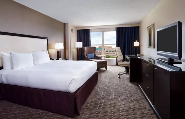 A modern hotel room with a large bed, white bedding, two bedside lamps, a TV on a dark dresser, a desk with a chair, an armchair by the window, and a city view outside.