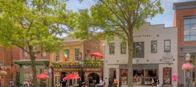A lively street scene with colorful historic buildings, outdoor seating with red umbrellas, green trees lining the sidewalk, and people dining and walking on a sunny day.