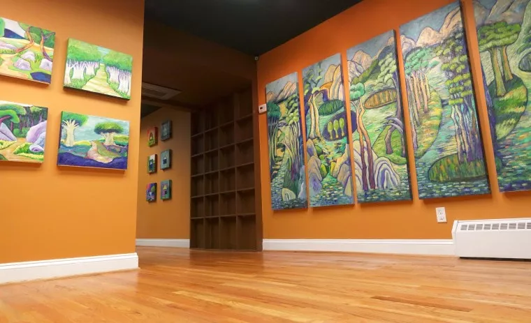 Art gallery with orange walls and wooden floors, displaying colorful landscape paintings, including a large multi-panel piece and smaller square artworks, with an empty wooden shelf in the background.