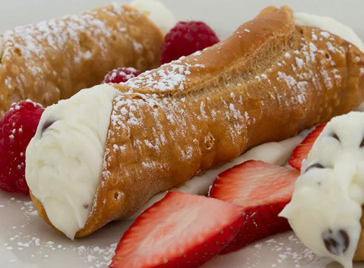 Two cannoli pastries filled with creamy ricotta filling, garnished with powdered sugar, fresh raspberries, and sliced strawberries on a light-colored plate.