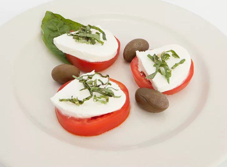 Three slices of tomato topped with mozzarella cheese and shredded basil, served with two black olives and a fresh basil leaf on a white plate.