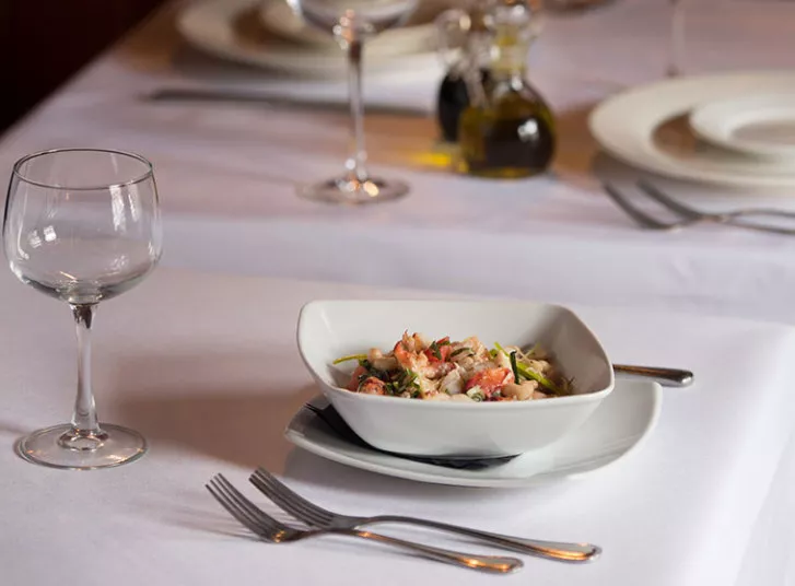 A white bowl of seafood salad sits on a white plate at a neatly set dining table with a wine glass, fork, knife, and spoon. Other plates and condiment bottles are in the softly blurred background.