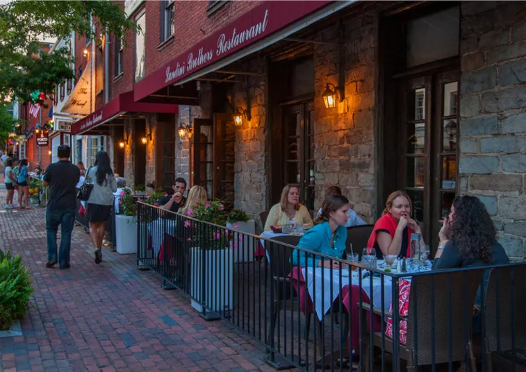 People dining at outdoor tables on a brick sidewalk in front of a restaurant with stone walls and red awnings on a lively street in the evening. Warm lights illuminate the scene, creating a cozy atmosphere.