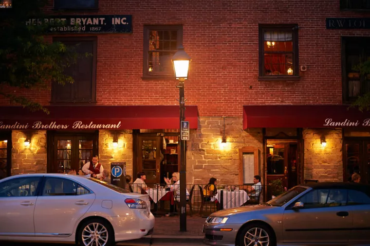 Outdoor diners sit at tables under red awnings at Landini Brothers Restaurant in the evening, with warm lights glowing and cars parked on the street in front of a brick building.