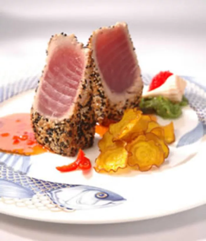 Two slices of seared, sesame-crusted tuna are plated upright with golden potato chips, colorful garnish, green wasabi, and a small pool of orange sauce on a decorative fish-patterned plate.