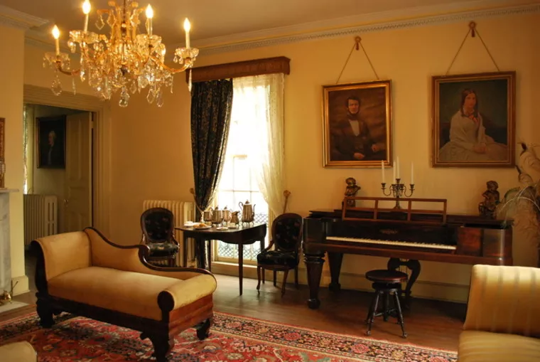 A vintage living room with a chandelier, antique furniture, a piano, ornate rug, large window with drapes, and two portraits hanging on the yellow walls above the piano.