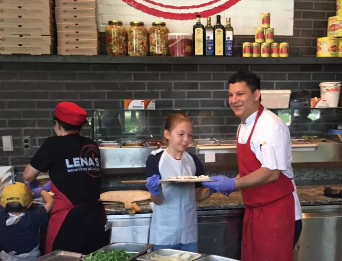 A chef and a young girl, both wearing aprons and gloves, smile while holding a pizza in a pizzeria kitchen. Another person prepares food nearby, with pizza ingredients and boxes in the background.