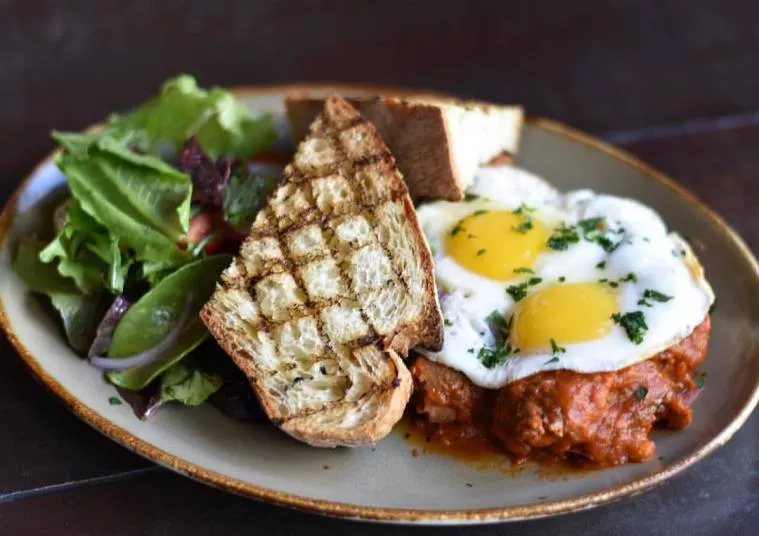A plate with two sunny-side-up eggs on a tomato-based sauce, two slices of grilled bread, and a side salad with mixed greens and red onion.