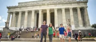 A family of four walks down the steps in front of the Lincoln Memorial in Washington, D.C., with groups of people scattered on the steps and near the large columns.