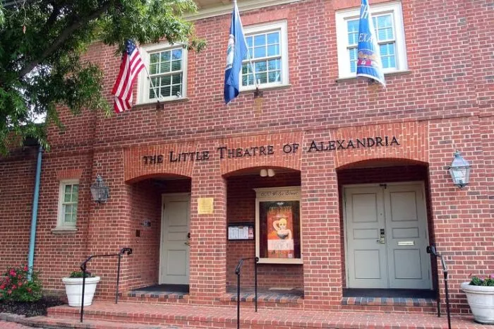 The brick exterior of The Little Theatre of Alexandria, with three flags above the entrance, two grey doors, potted plants, and a poster displayed between the doors.