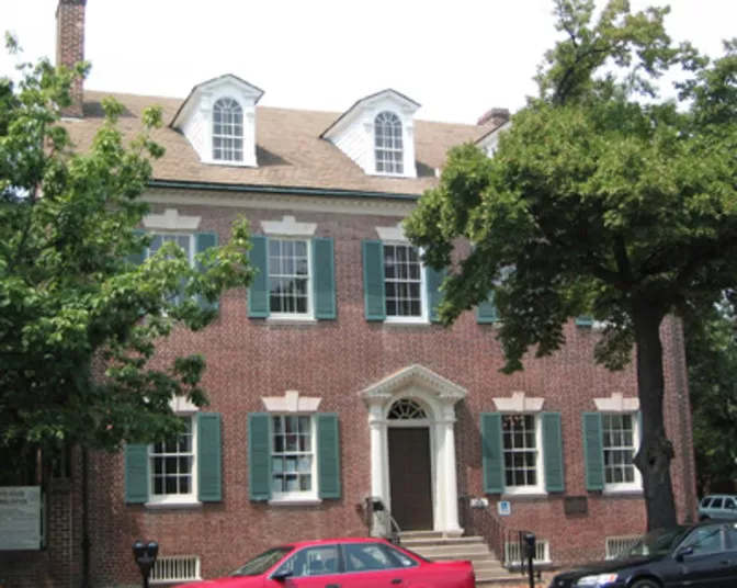 A large, historic brick building with green shutters, white trim, dormer windows, and an arched doorway, partially shaded by trees. Several parked cars are visible in front.