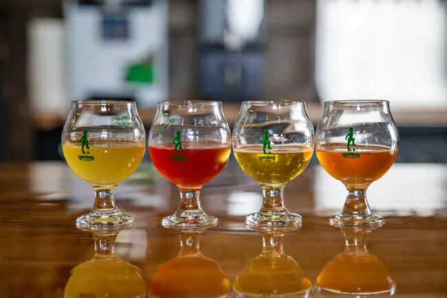 Four stemmed glasses filled with different colored beverages—yellow, red, pale yellow, and orange—are lined up on a shiny wooden table with a blurred background.