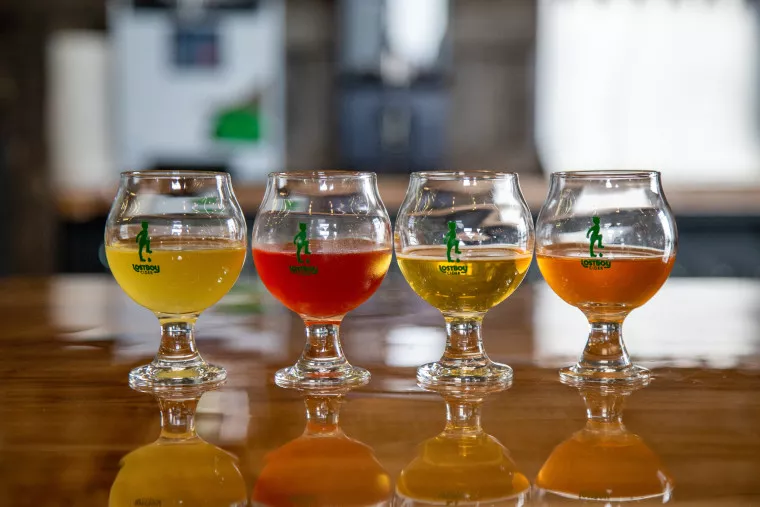 Four stemmed glasses filled with different colored beverages—yellow, red, pale yellow, and orange—are lined up on a shiny wooden table with a blurred background.