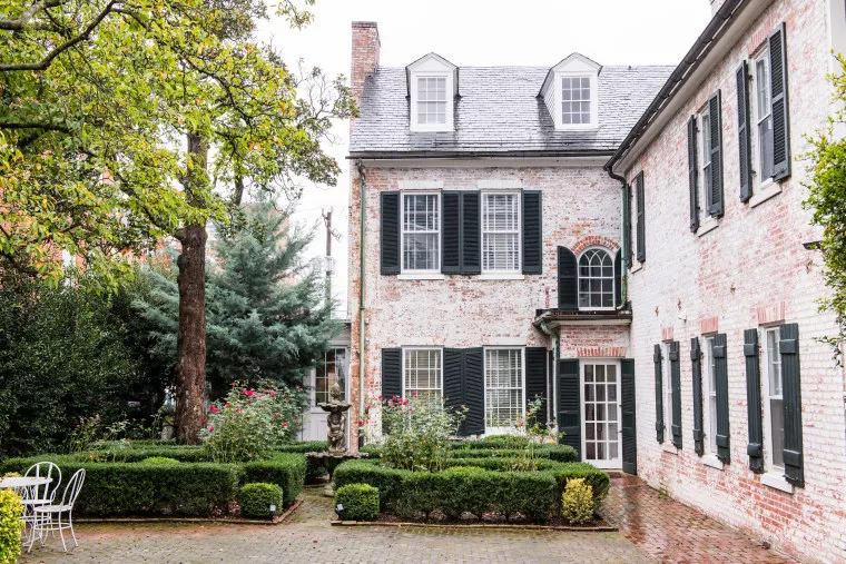 A brick courtyard features green hedges, a small fountain, a white metal table with chairs, and two historic brick buildings with dark green shutters and multiple windows.