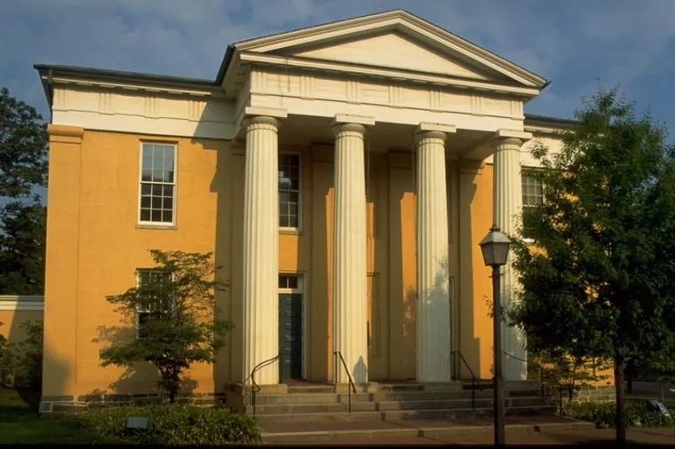 A yellow neoclassical building with four tall white columns at the entrance, wide steps, and a triangular pediment. Green trees and a black lamp post stand in front, under a blue sky with some clouds.