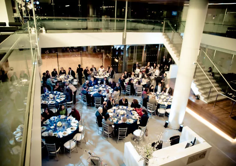A large group of people seated at round tables, dressed formally, attend a dinner event in a modern, open space with glass railings and a staircase.