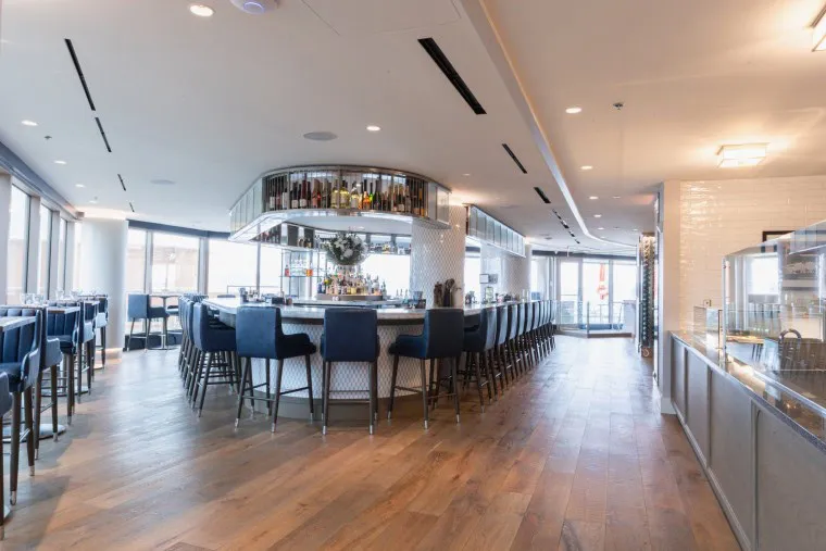 Modern, elegant restaurant interior featuring a central circular bar with blue barstools, wooden flooring, large windows with natural light, and a stylish, open atmosphere.