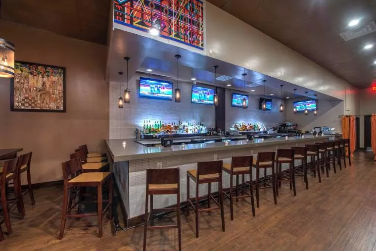 Modern bar with wooden floors, high chairs lined along a counter, shelves of liquor, and several TVs mounted above the bar. Colorful abstract artwork decorates the walls, and the space has warm lighting.