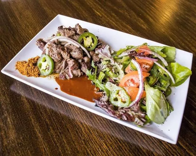 A rectangular white plate with grilled beef strips topped with sliced jalapeños and onions, served alongside mixed green salad with tomato slices, onions, and jalapeños. A small pile of spice and sauce are on the side.