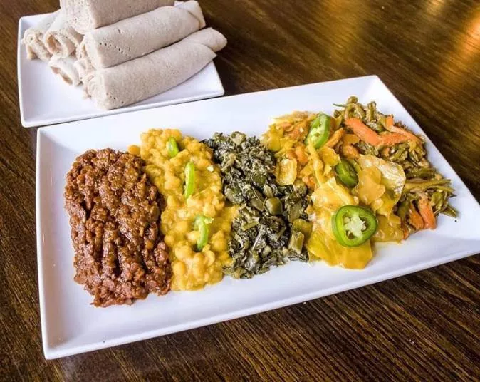 A rectangular white plate with four portions of colorful Ethiopian dishes, including stewed lentils, yellow split peas, greens, and mixed vegetables, with rolled injera bread on a separate plate in the background.