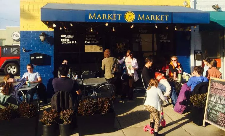People sit and socialize at outdoor tables in front of a café called Market Market. Some are eating and drinking, while children play nearby. The scene is lively and sunny, with potted plants lining the sidewalk.
