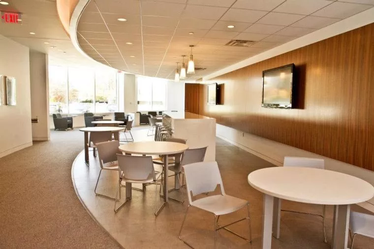 Modern office lounge area with round white tables, white chairs, a curved ceiling, pendant lights, and wall-mounted TVs. Large windows let in natural light. The space is clean and organized with minimal decor.