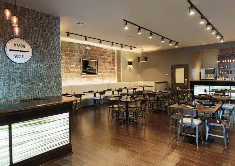 Modern restaurant interior with wooden floors, neatly arranged tables and chairs, a patterned accent wall with “Mason Social” signage, exposed brick, and a softly lit bar area in the background.