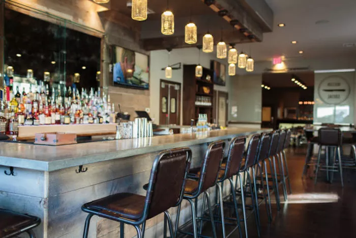 A modern bar with a long concrete countertop, high-back leather stools, hanging jar lights, and shelves of liquor bottles behind the counter. The space has a rustic, cozy ambiance with wood accents and warm lighting.