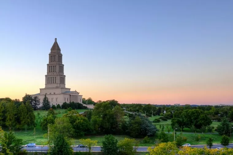 A tall, tiered stone building with a spire stands among green trees and lawns at sunset, with a clear sky and a few cars on a nearby road in the foreground.