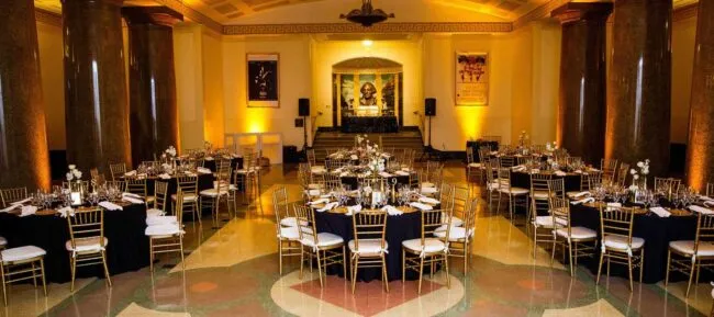 A grand banquet hall with round tables covered in black tablecloths and gold chairs, elegantly set for an event. Warm lighting highlights tall columns, ornate decor, and a statue at the far end of the room.