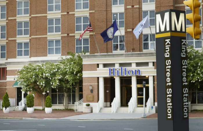 A Hilton hotel building with flags flying above the entrance is seen behind a King Street Station metro sign and traffic light on a city street.