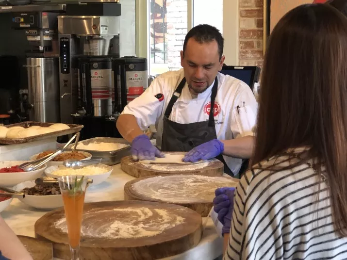 A chef in a white uniform and purple gloves spreads flour on wooden pizza boards, preparing dough as people watch. Ingredients and a drink are visible on the counter beside him.