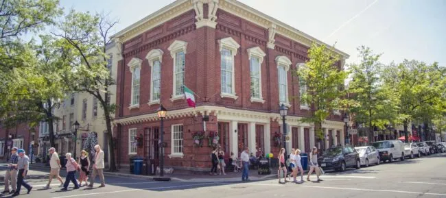 A large red-brick building with tall windows and an Italian flag on the corner, surrounded by trees and people walking on a sunny day. Several cars are parked along the street.