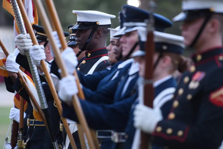 A row of uniformed military personnel from different branches stand side by side, holding flags and rifles during a ceremonial event outdoors.