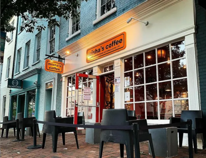 Outdoor view of Misha’s Coffee shop with large windows, orange sign, empty black chairs and tables on a brick sidewalk. The street has colorful buildings and tree branches overhead.