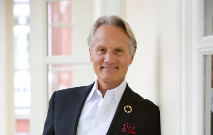 A smiling man with gray hair wears a black suit jacket, white dress shirt, and a decorative brooch on his lapel, standing indoors near a window.