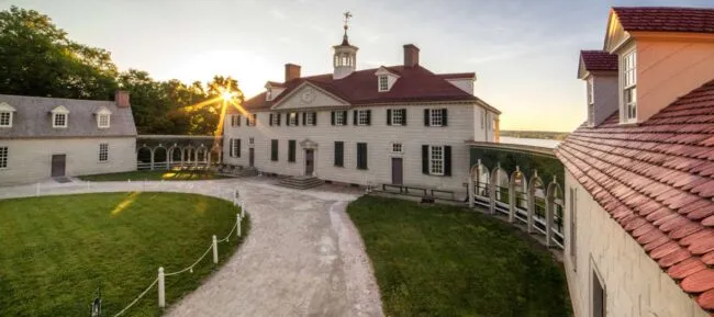 A wide-angle view of a historic mansion with white walls, green shutters, and a red roof, surrounded by green lawn, a gravel path, and a colonnade, seen at sunrise or sunset.