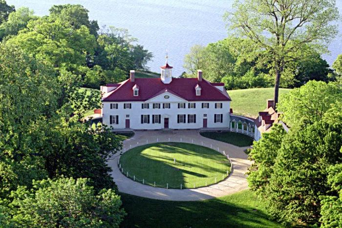 A large historic white house with a red roof sits at the center of a circular driveway, surrounded by green trees and lawn, with water visible in the background.