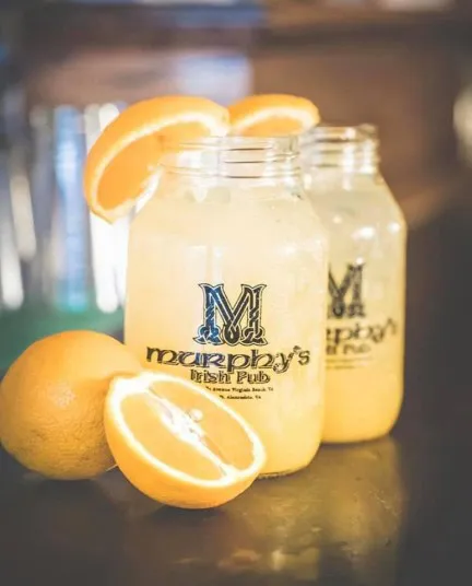 Two mason jars filled with light yellow lemonade, garnished with orange slices, sit on a counter beside a halved orange. The jars are labeled Murphys Irish Pub in black text.