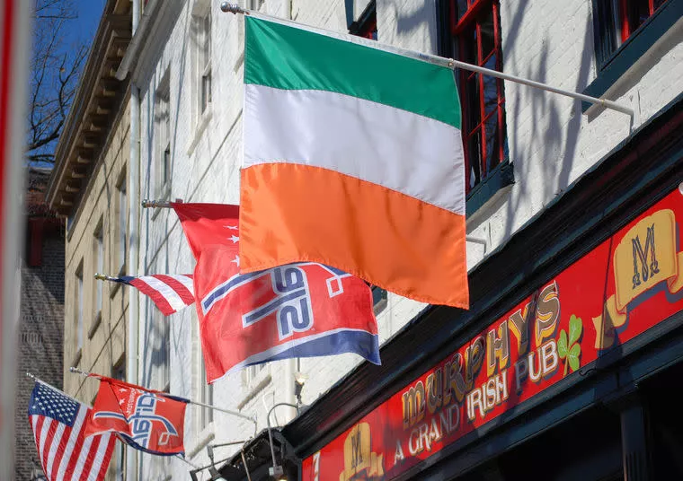 Three flags—Irish, Washington Capitals, and American—hang outside Murphy’s Grand Irish Pub on a sunny day, with the building’s sign partially visible and red window frames above.