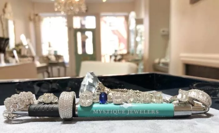 A collection of diamond and gemstone rings and bracelets displayed on a turquoise pen labeled MYSTIQUE JEWELERS, with a blurred view of a jewelry store interior in the background.